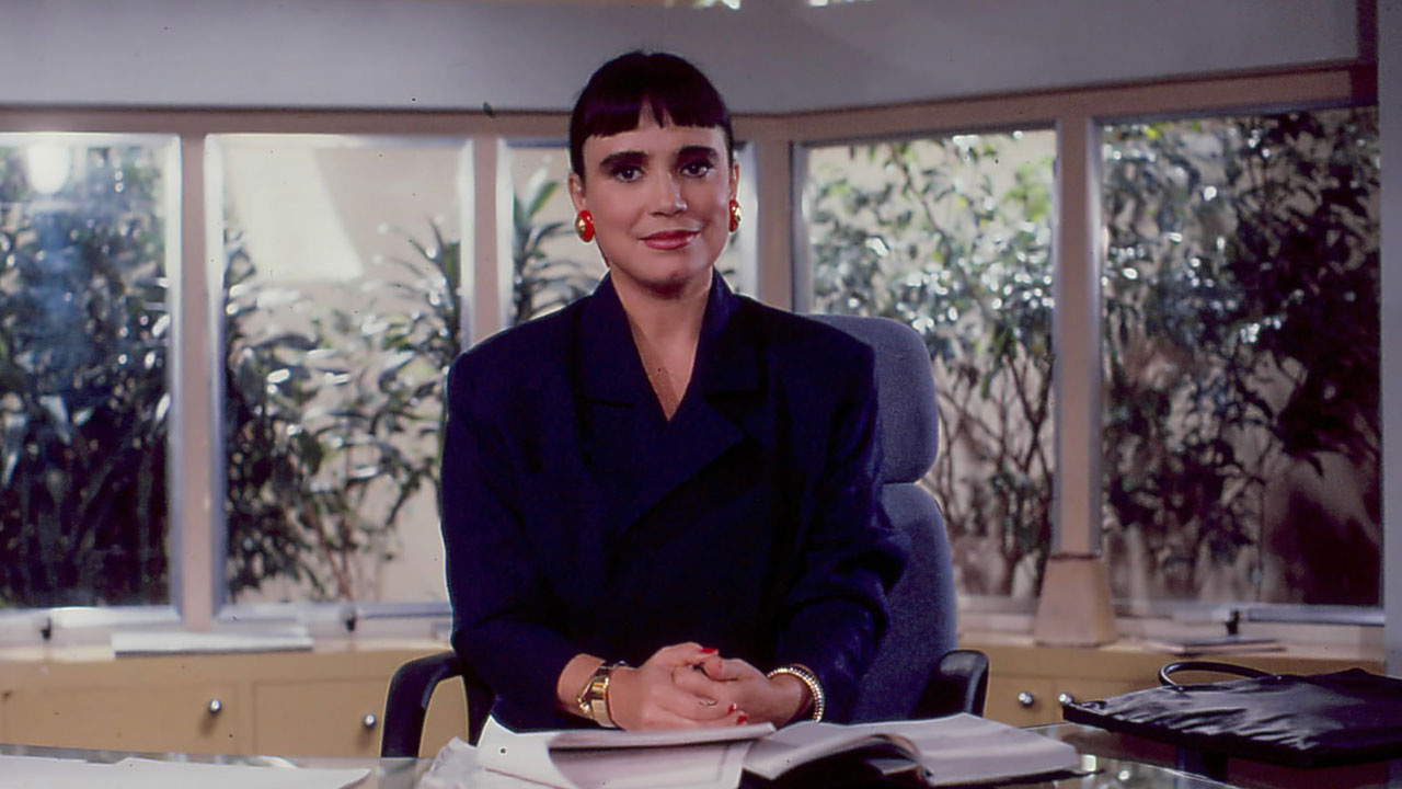 Regina Duarte como Maria do Carmo com seu clássico cabelo de franja, sentada com as mãos juntas na mesa de seu escritório em Rainha da Sucata
