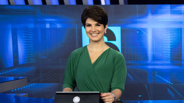 Mariana Godoy, de blusa verde e cabelo curto, sorri em pé atrás da bancada de um moderno estúdio de telejornal