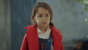 Melek Akcay, interpretada por Beren Gokyildiz, usando casaco vermelho sobre uniforme escolar em cena da novela Mãe