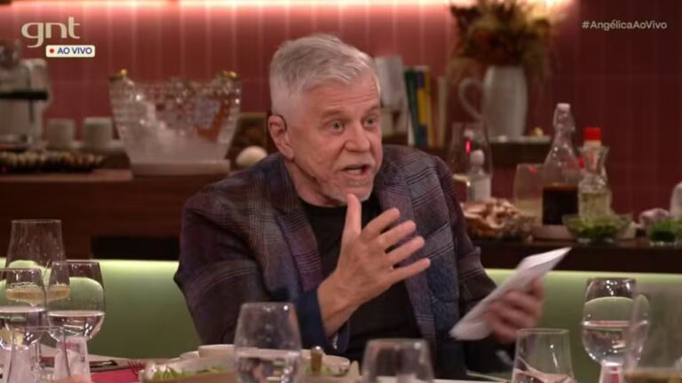 Miguel Falabella durante uma conversa em programa de TV, gesticulando com uma das mãos enquanto segura um papel, em mesa posta com taças e pratos, em ambiente elegante do canal GNT
