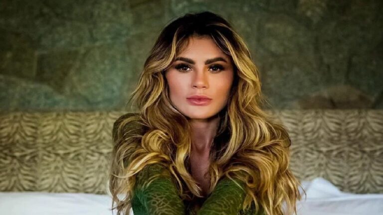 Natacha Horana posa sentada, com expressão séria e cabelos loiros soltos, usando blusa verde e fundo neutro desfocado