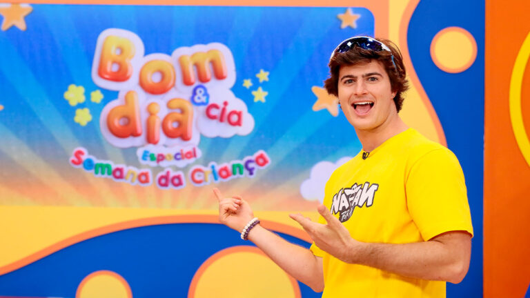 Natan Por Aí aponta para o logotipo do Bom dia & Cia Especial Semana da Criança no estúdio do programa infantil do SBT