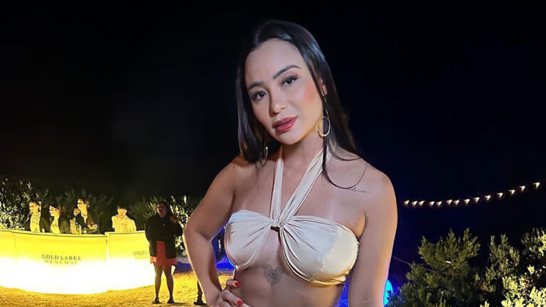 Pablinny Pedersoli com top bege e cabelos escuros posa para foto durante um evento noturno com um bar iluminado ao fundo