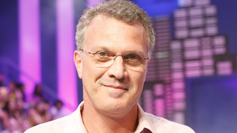 Pedro Bial durante período em que apresentava o Big Brother Brasil com expressão sorridente e cenário colorido ao fundo