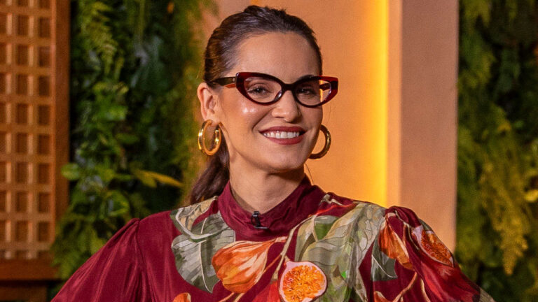 A chef Renata Vanzetto sorri com óculos de armação gatinho, argolas douradas e blusa vermelha com estampa de folhas e figos