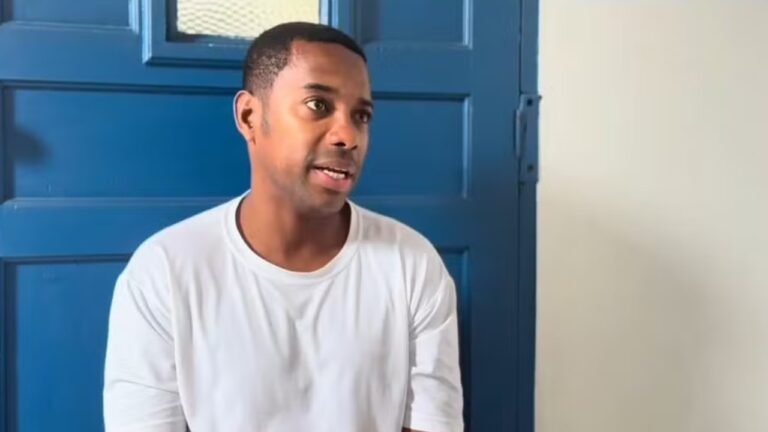 Ex-jogador Robinho sentado de camiseta branca fala em entrevista com porta azul ao fundo