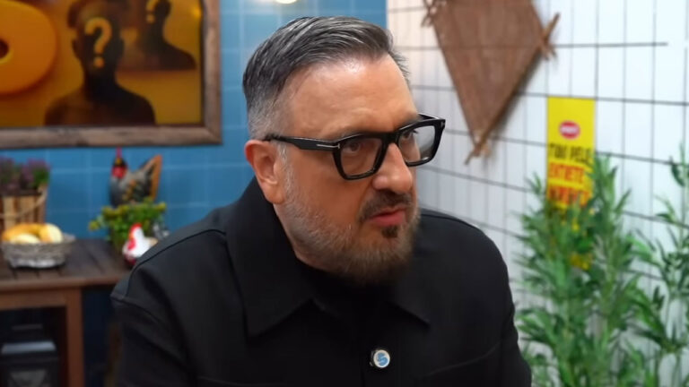 O diretor Rodrigo Carelli, de óculos e jaqueta preta, fala com expressão séria durante entrevista em um estúdio de gravação