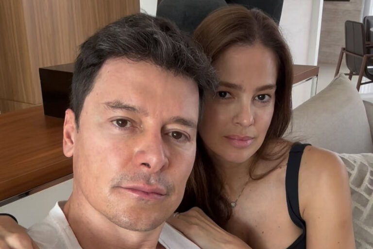 Homem e mulher sentados juntos em sofá de sala moderna posam para selfie com expressões sérias e olhar voltado para a câmera