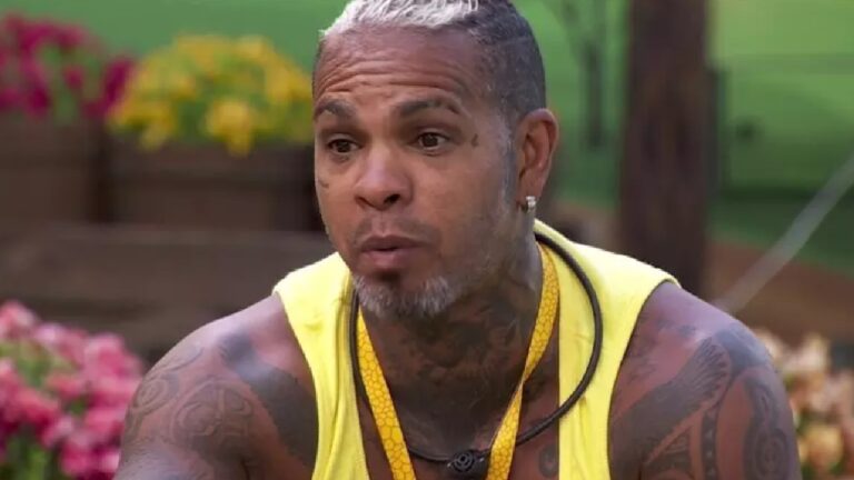 Rodriguinho em um momento de conversa ao ar livre, usando regata amarela e colar preto, com expressão séria e flores coloridas ao fundo