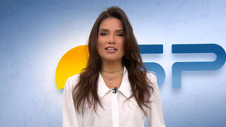 A jornalista Sabina Simonato, de camisa branca e colares, apresenta telejornal local no estúdio da Globo com o logo SP ao fundo
