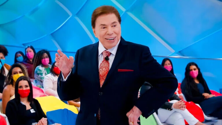 Silvio Santos com terno azul e gravata vermelha sorri no estúdio do SBT durante gravação de seu programa com plateia usando máscaras