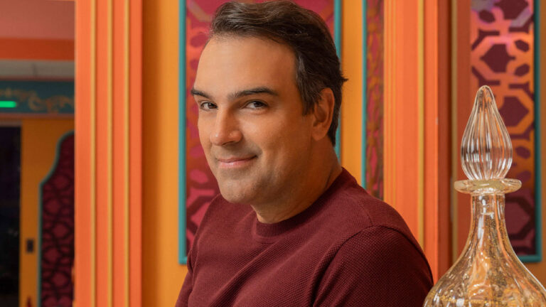 Tadeu Schmidt sorrindo em enquadramento fechado de suéter vinho com fundo de listras coloridas do BBB 25