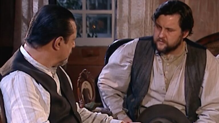 Cena da novela Terra Nostra, mostrando dois homens em trajes do século XIX sentados e conversando em uma sala com iluminação suave e decoração de época, reforçando o clima histórico do drama