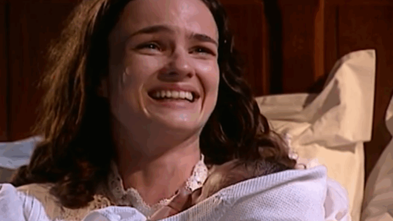 Cena da novela Terra Nostra mostra uma mulher emocionada e sorrindo, deitada em uma cama com lençóis brancos, segurando um bebê recém-nascido nos braços