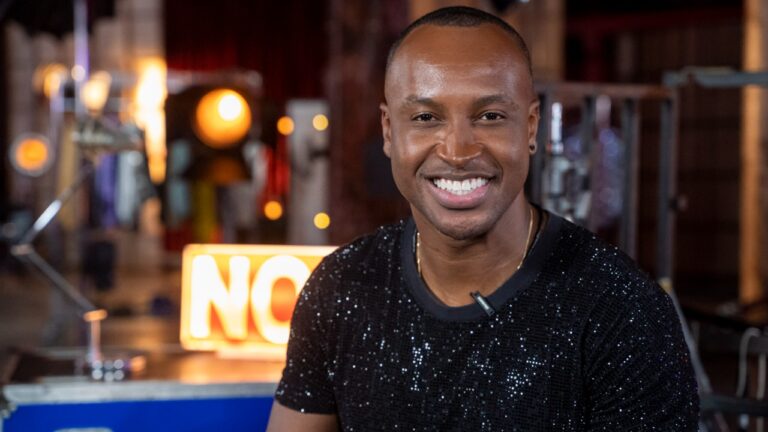 Thiaguinho sorrindo com uma camiseta preta com brilho em estúdio iluminado com refletores e letreiro luminoso desfocado ao fundo