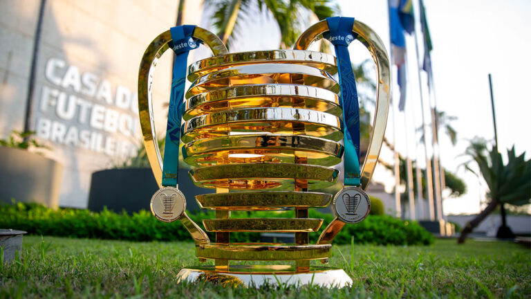 Troféu da Copa do Nordeste dourado com alças e anéis horizontais sobre a grama com bandeiras ao fundo