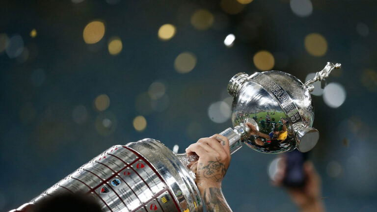 Mão de jogador com tatuagens ergue o troféu da Taça Libertadores em meio a comemoração com chuva de papel picado no estádio