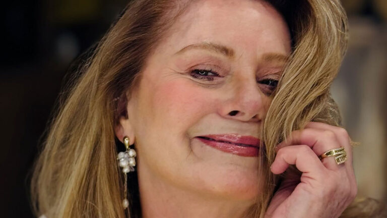 Vera Fischer sorrindo, com cabelo loiro e a mão esquerda perto do rosto. Ela usa um brinco longo de pérolas e um anel