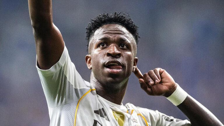 O jogador Vini Jr comemorando um gol com a mão esquerda na orelha e o braço direito erguido vestindo a camisa do Real Madrid