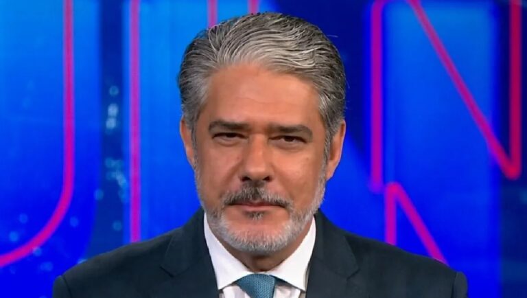 William Bonner de camisa branca e gravata azul enquanto olha para a câmera em estúdio de telejornal com fundo azul iluminado por luzes coloridas e letras desfocadas