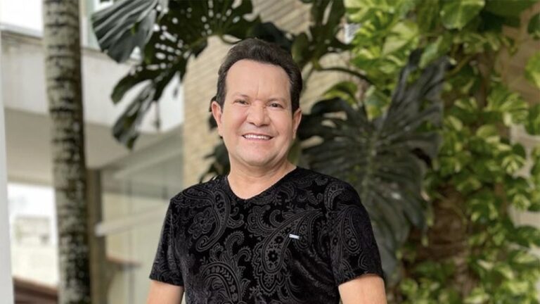 Ximbinha sorrindo usa camiseta preta com estampa trabalhada posando em área externa com plantas grandes ao fundo