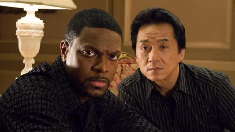 Dois homens sentados olhando seriamente para frente em ambiente interno iluminado por abajur