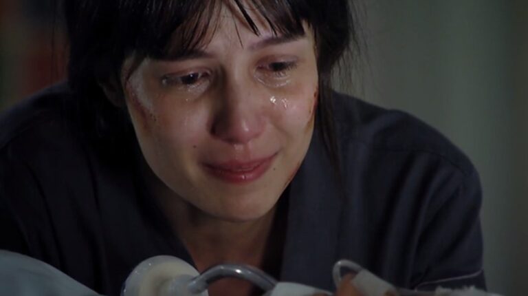 A imagem mostra Marjorie Estiano em A Vida da Gente chorando intensamente, com lágrimas escorrendo pelo rosto e expressão de sofrimento. Ela parece estar em um ambiente fechado, possivelmente um hospital ou local de tensão emocional