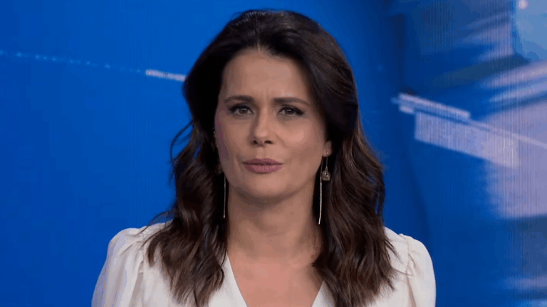 Adriana Araújo com blusa branca e brincos longos, em plano médio, apresentando o Jornal da Band em estúdio azul
