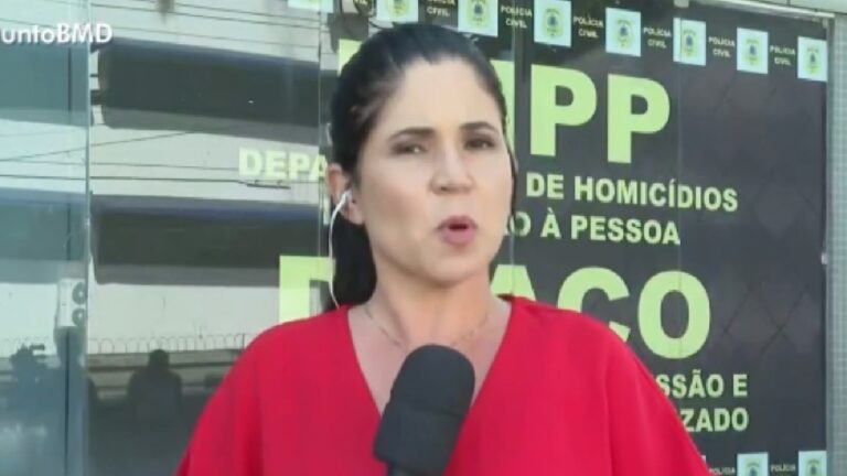 Repórter veste blusa vermelha e segura microfone enquanto faz entrada ao vivo em frente a uma delegacia, com placas da Polícia Civil visíveis ao fundo