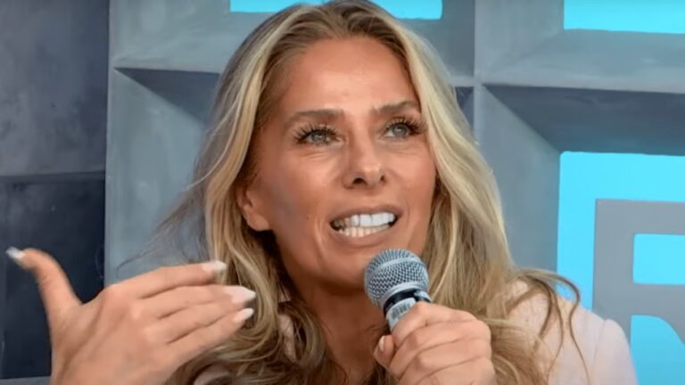 Mulher loira segura microfone e fala sorrindo durante entrevista em ambiente iluminado