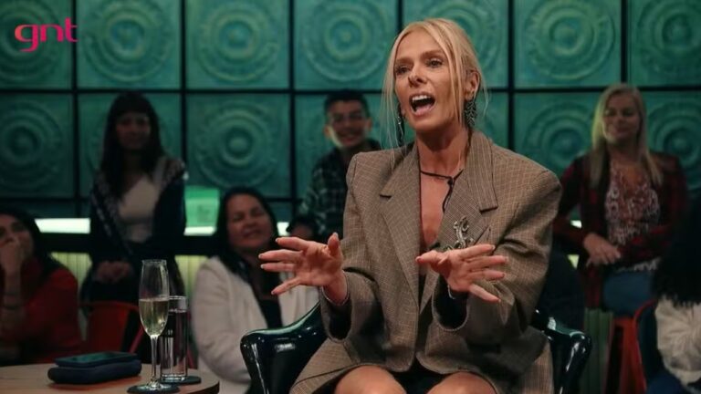 Mulher loira fala animadamente durante programa de TV do canal GNT, sentada diante de plateia, usando blazer xadrez e com taça sobre a mesa à frente