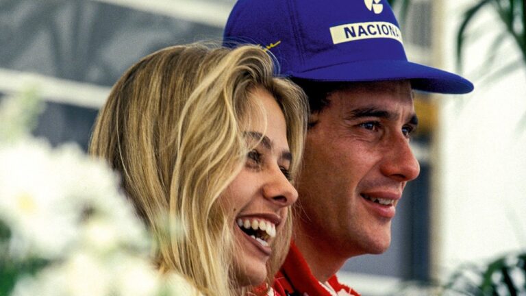 Homem com boné azul e mulher loira sorriem lado a lado em evento esportivo