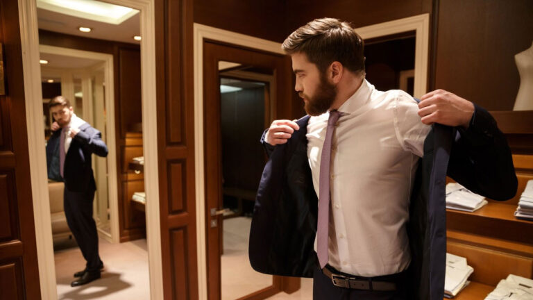 Homem de barba vestindo um paletó azul-marinho, camisa social branca e gravata rosa em frente a um espelho em uma loja