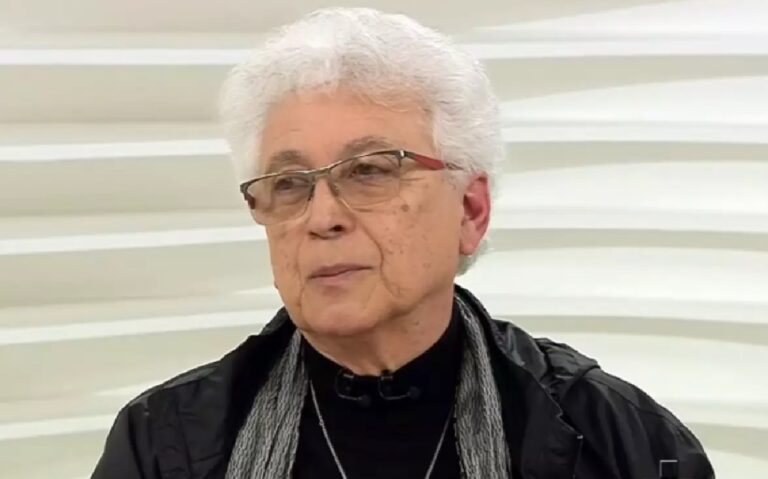 A imagem mostra uma pessoa idosa de cabelos brancos curtos e óculos, usando uma jaqueta preta, cachecol cinza e uma blusa escura. O fundo é claro, com um padrão ondulado branco, típico de um estúdio de televisão