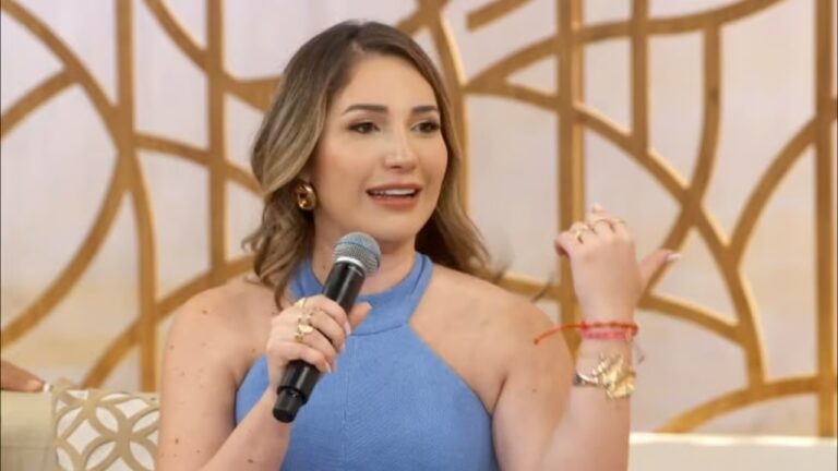 A imagem mostra uma mulher falando em um programa de TV ou entrevista, segurando um microfone enquanto gesticula com a outra mão. Ela veste uma blusa azul sem mangas e usa diversos acessórios dourados, incluindo brincos, anéis e uma pulseira, além de uma fita vermelha no pulso. A mulher está sentada em um ambiente claro e elegante, com um painel decorativo geométrico ao fundo. Ela sorri levemente, transmitindo simpatia e naturalidade durante a conversa