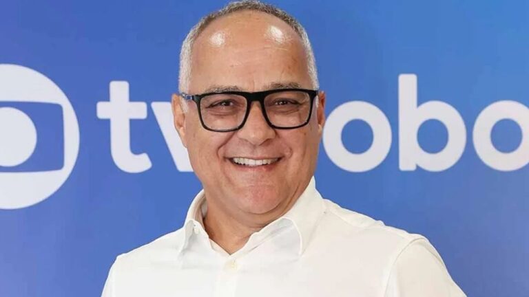 Homem sorridente posa diante de um painel azul com o logotipo de uma emissora de TV ao fundo. Ele usa camisa social branca e óculos de armação preta, transmitindo postura profissional em ambiente de divulgação institucional