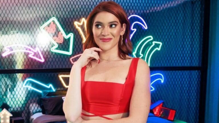 Mulher de cabelos ruivos sorri com expressão confiante, vestindo top vermelho, em ambiente moderno decorado com setas de luz neon coloridas ao fundo