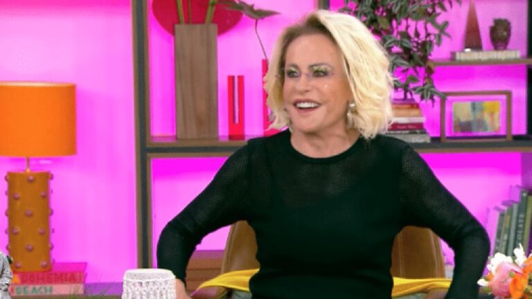 Mulher de cabelos loiros curtos sorri enquanto participa de um programa de TV em estúdio colorido. Ela veste blusa preta de tecido vazado e está sentada em uma poltrona, com as mãos apoiadas nos braços. Ao fundo, há prateleiras com livros, plantas e objetos decorativos sobre uma parede iluminada em rosa vibrante.