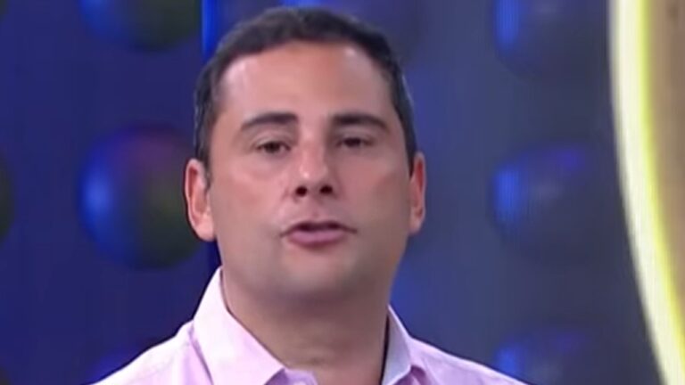 A imagem mostra um homem com expressão séria, vestindo uma camisa social rosa-clara. O fundo é azul com elementos circulares desfocados, sugerindo que ele está em um estúdio de televisão.