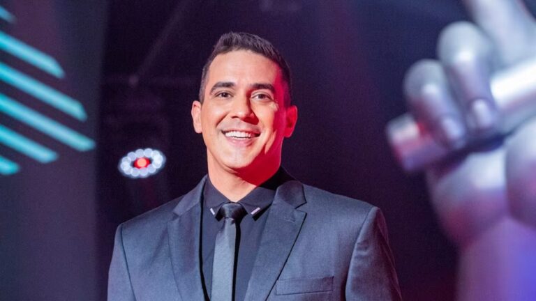 André Marques de terno e gravata sorri em um estúdio iluminado, com cenário moderno e uma grande escultura de mão ao fundo, sugerindo ambiente de programa de televisão