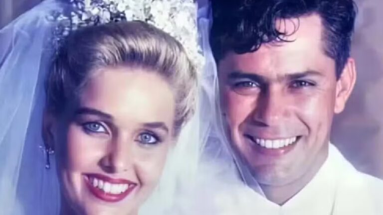 A imagem mostra um casal sorridente vestido para um casamento. A mulher usa um vestido de noiva branco com véu e uma tiara adornada com flores, enquanto o homem está com um terno branco. Ambos estão muito próximos e olhando diretamente para a câmera, transmitindo felicidade e um clima de celebração