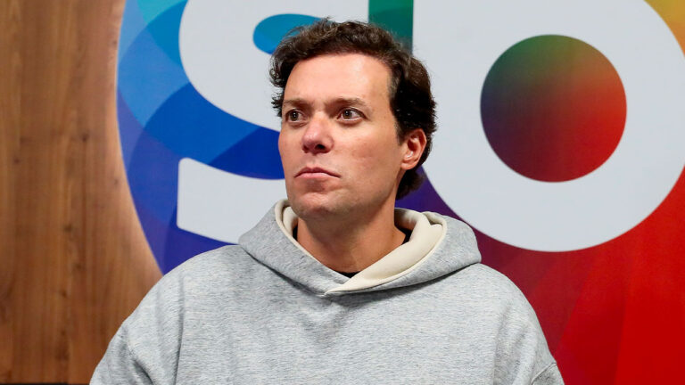 André Valadão de moletom cinza e capuz, em plano médio, olhando para o lado no fundo com o logo do SBT