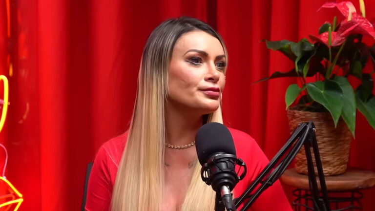 Andressa Urach com cabelos loiros longos, vestindo blusa vermelha e falando em um microfone de estúdio de podcast