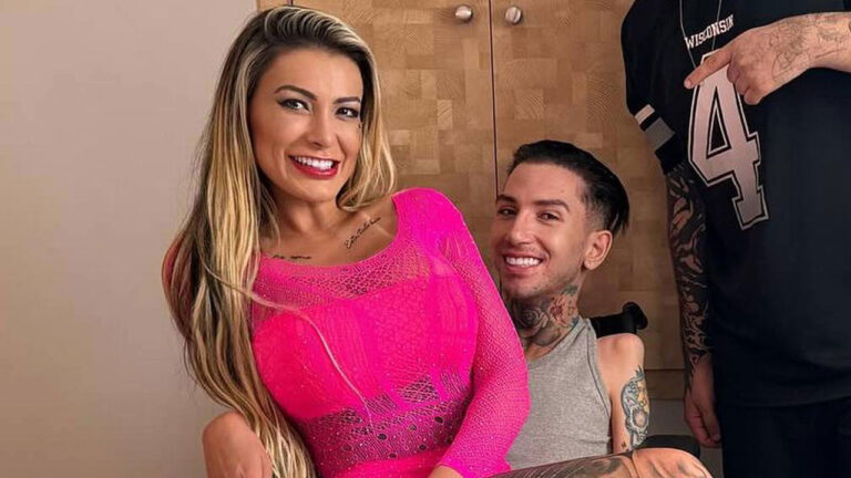 Andressa Urach com roupa pink de tela e Gabriel Brito, de regata cinza, sorriem em foto em ambiente interno