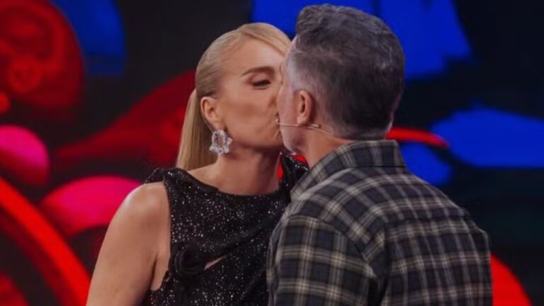 Casal se beija no palco de programa de televisão com cenário colorido ao fundo