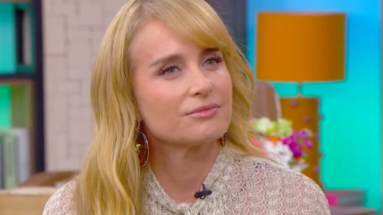 A imagem mostra uma mulher loira de cabelos compridos, vestindo uma blusa clara rendada. Ela está em um estúdio colorido, com uma luminária laranja e flores ao fundo