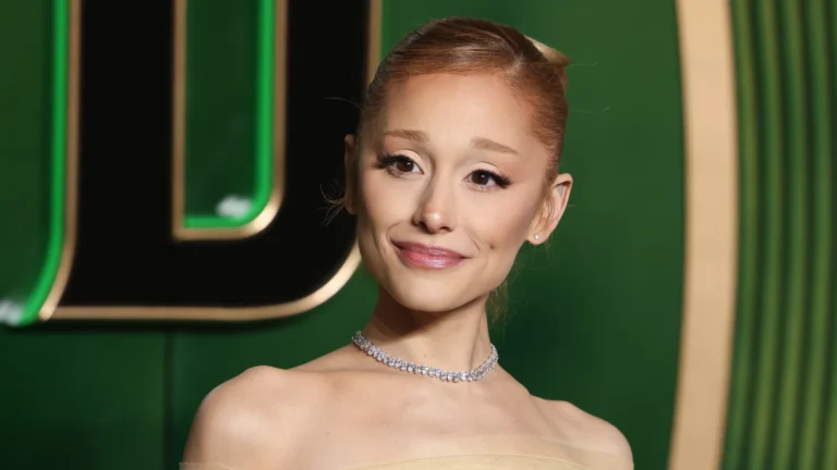 Mulher em evento de tapete vermelho posa diante de um fundo verde com detalhes dourados. Ela usa vestido tomara que caia em tom claro, colar brilhante de pedras e brincos pequenos. O cabelo está preso em um coque elegante, e a maquiagem destaca olhos e lábios com acabamento suave