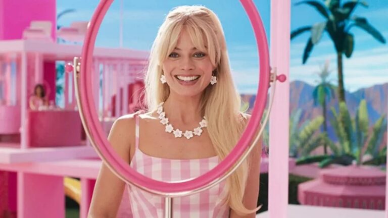 Mulher loira sorri em frente a um espelho circular rosa em um cenário vibrante e inteiramente cor-de-rosa, com arquitetura estilizada e palmeiras ao fundo. Ela usa vestido xadrez rosa e branco, além de colar e brincos de flores brancas, transmitindo estética lúdica e decorativa