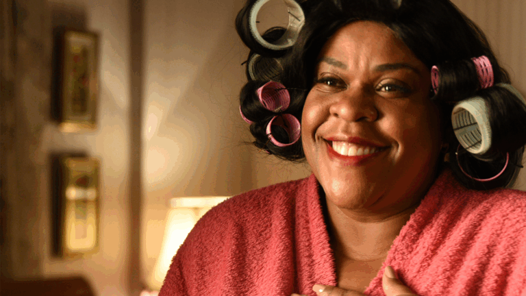 A imagem mostra uma mulher sorridente, vestindo um roupão rosa e com vários bobes coloridos (rosas e verdes) no cabelo. Ela parece estar em um ambiente doméstico, com uma iluminação quente e suave, transmitindo uma sensação de conforto e bom humor. Ao fundo, é possível ver quadros pendurados e um abajur aceso