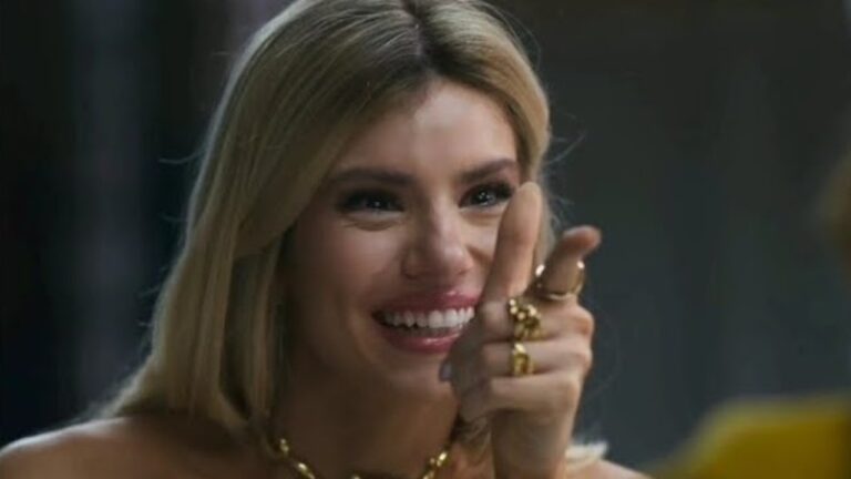 Mulher sorridente, com cabelos loiros soltos e acessórios dourados, aponta para frente de forma descontraída, exibindo anéis e colar enquanto olha para alguém fora do quadro